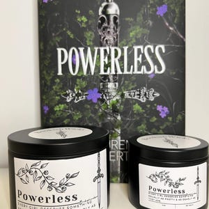 May include: Two black candle tins with white labels. The labels feature a sword and a vine design. The text on the labels reads "Powerless" and "Every girl deserves something equally as pretty & as deadly as they are."