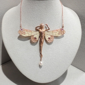 Könnte beinhalten: Halskette aus Roségold mit einem Anhänger in Form einer stilisierten Fee. Die Flügel sind mit cremefarbenem und rosa Emaille verziert und mit kleinen roten Edelsteinen versehen. Eine einzelne Perle hängt von der Fee herab.