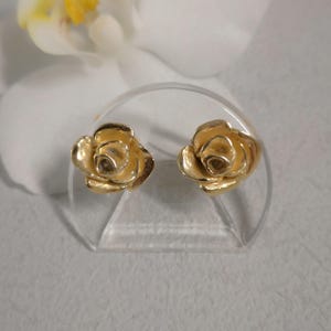 Op de afbeelding: Een paar goudkleurige roosvormige oorstekers. De oorbellen worden gepresenteerd op een heldere, gebogen standaard, met een witte bloem op de achtergrond. De oorbellen hebben gedetailleerde bloemblaadjes en een glanzende afwerking.