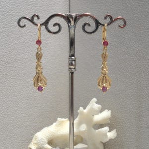 Op de afbeelding: Een paar gouden oorhangers met een delicaat, versierd ontwerp. Elke oorbel heeft bovenaan en onderaan een kleine, ronde, roze edelsteen.