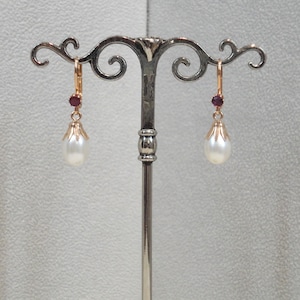 Op de afbeelding: Een paar gouden oorhangers met een witte parel en een kleine rode edelsteen. De oorbellen hangen aan een zilveren standaard.