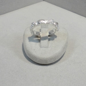 Könnte beinhalten: Ein silberner Ring mit einem strukturierten Design aus Muscheln. Der Ring wird auf einem weißen Ringständer präsentiert.