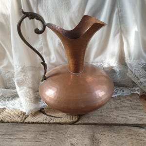 May include: A hammered copper jug with a decorative handle. The jug has a wide mouth and a rounded body.