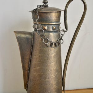 May include: A vintage brass teapot with a hammered finish and a chain handle. The teapot has a spout and a lid with a decorative knob. The teapot is sitting on a wooden surface.