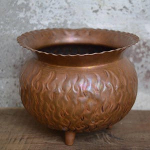 Puede incluir: Un macetero de cobre con un patrón de llama y tres patas. El macetero está hecho de metal martillado y tiene un aspecto rústico y vintage.