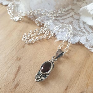 Può includere: Una collana con catena d'argento con un ciondolo che presenta una pietra ovale di colore rosso scuro incastonata in una cornice argentata ornata.