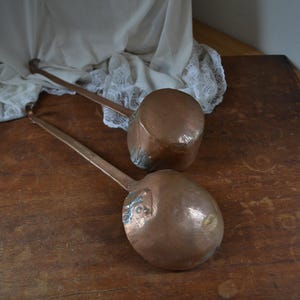 Könnte beinhalten: Zwei antike Kupferlöffel mit langen Holzgriffen. Die Löffel liegen auf einer Holzoberfläche.