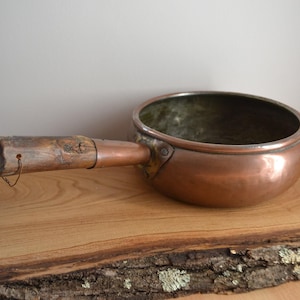 May include: A vintage copper pot with a wooden handle. The pot has a hammered finish and is sitting on a wooden surface.