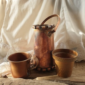 May include: Three copper mugs, one with a handle, on a wooden surface. The mugs are all different sizes and shapes.