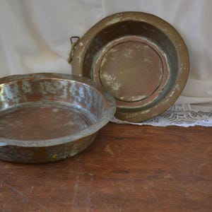 May include: Two antique copper bowls with a worn, aged patina. The bowls are round and have a slightly raised rim. The larger bowl has a handle on the side.