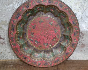 May include: A round, decorative brass plate with a scalloped edge. The plate is painted with intricate floral designs in shades of red, green, and gold.