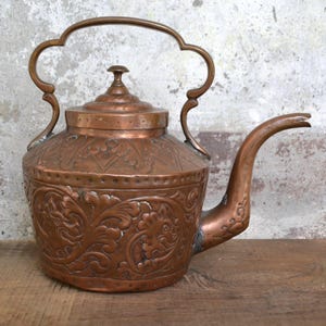 Puede incluir: Una tetera de cobre antigua con un diseño floral ornamentado. La tetera tiene una tapa y un pico, y el asa tiene forma de bucle.