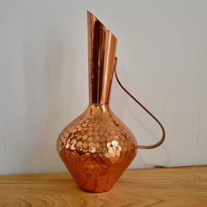 May include: A copper jug with a hammered texture on the rounded base and a smooth, elongated neck. The jug has a handle and an angled spout. The jug is on a wooden surface.