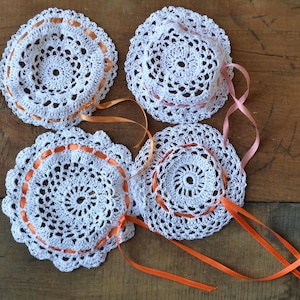 Peut inclure: Quatre napperons en crochet blanc avec une bordure de ruban orange. Les napperons sont disposés sur une surface en bois.