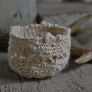 May include: A small, white crocheted basket with a delicate, lacy design. The basket is made of natural fibers and has a soft, textured surface.