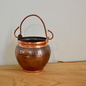May include: A small copper pot with a handle. The pot has a hammered finish and is sitting on a wooden surface.