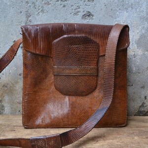 Può includere: Una borsa a tracolla in pelle marrone con chiusura a lembo e una tasca più piccola sul davanti. La borsa ha un motivo testurizzato, simile a quello della pelle di serpente.