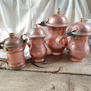 May include: Four copper teapots with lids and handles. The teapots are arranged on a wooden surface. The teapots are all different sizes and shapes.