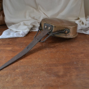 May include: An antique copper saucepan with a long, riveted iron handle. The pan has a rounded base and a small loop at the end of the handle. The handle extends approximately 46 cm from the pan. The pan is set on a wooden surface.