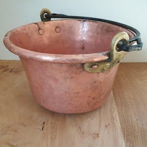 May include: A copper pot with a black iron handle and brass accents. The pot has a rounded bottom and a wide opening. It is made of hammered copper and has a rustic look.