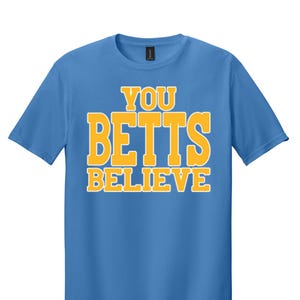 May include: A light blue t-shirt with the text "YOU BETTS BELIEVE" in yellow.