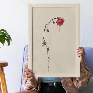 Puede incluir: Impresión en acuarela enmarcada con una rosa roja con tallo y hojas caídas. La obra de arte tiene una estética vintage, con gotas rojas que se extienden desde la flor. El marco es de color beige claro.