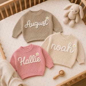 May include: Three long-sleeve knit jumpers in beige, cream, and pink. Each jumper features a white cursive name embroidered on the front: August, noah, and Hallie. A small white daisy applique with a yellow centre is attached next to each name.