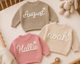 Op de afbeelding: Drie gebreide truien met lange mouwen in beige, crème en roze. Elke trui heeft een witte, in cursief geborduurde naam op de voorzijde: August, noah en Hallie. Naast elke naam is een kleine witte madeliefjesapplicatie met een geel hartje bevestigd.