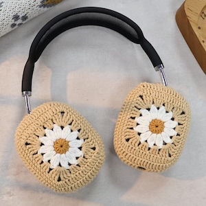 Puede incluir: Fundas para auriculares de crochet beige con un dise&ntilde;o de margarita blanca. Las fundas est&aacute;n unidas a una diadema negra.