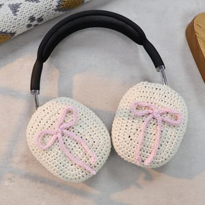 Pu&ograve; includere: Paraorecchie in crochet bianco con fiocchi rosa. I paraorecchie sono attaccati a una fascia nera.