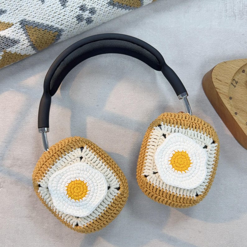 May include: A pair of crocheted headphone covers in the shape of a fried egg. The covers are beige with a white egg yolk and a yellow yolk.