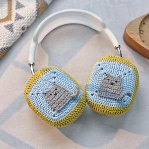 Pu&ograve; includere: Cuffie bianche con paraorecchie all'uncinetto con un design di gatto grigio. I paraorecchie sono gialli e blu.