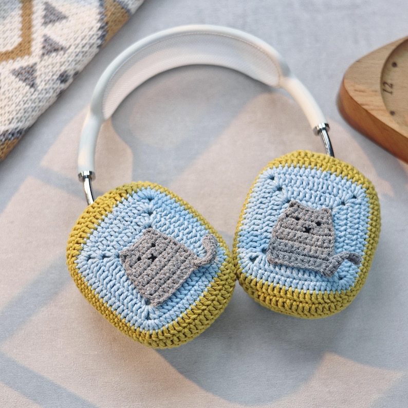 Pu&ograve; includere: Cuffie bianche con paraorecchie all'uncinetto con un design di gatto grigio. I paraorecchie sono gialli e blu.