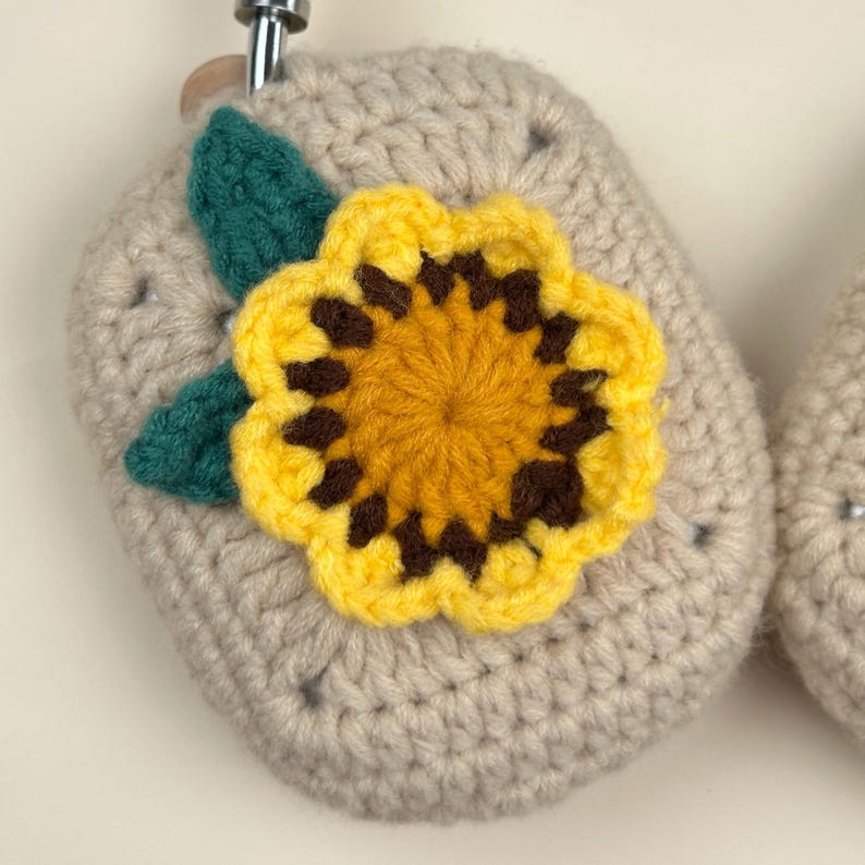 Op de afbeelding: Gehaakte beige vierkant met een gele en bruine zonnebloem en een groen blad. De zonnebloem is gecentreerd op het vierkant. Het item heeft een metalen lus aan de bovenkant.