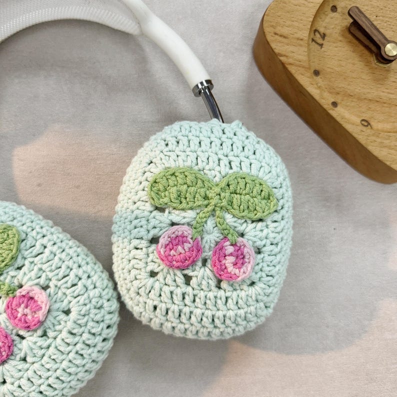 Pu&ograve; includere: Un paio di copri cuffie all'uncinetto di colore verde chiaro con un design a ciliegie. Le ciliegie sono rosa e bianche e hanno foglie verdi.