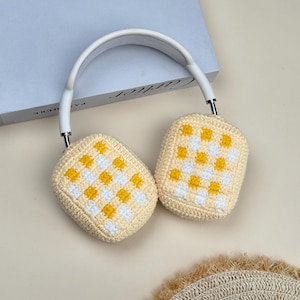 May include: White and silver headphones with cream-colored, crocheted ear covers. The ear covers feature a yellow and white checkered pattern. The headphones are resting on a book.
