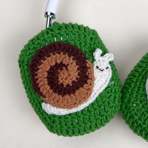Pu&ograve; includere: Copri cuffie all'uncinetto verdi con un design a chiocciola marrone e beige. La chiocciola ha un corpo bianco e un viso sorridente. Le cuffie sono bianche.
