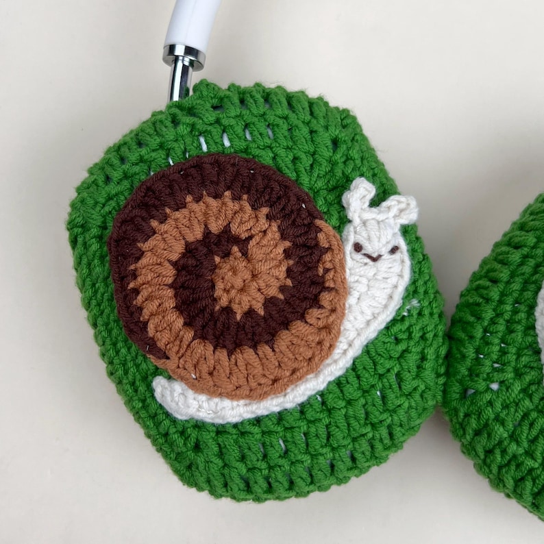 Pu&ograve; includere: Copri cuffie all'uncinetto verdi con un design a chiocciola marrone e beige. La chiocciola ha un corpo bianco e un viso sorridente. Le cuffie sono bianche.