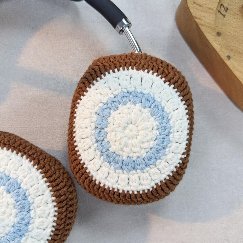 May include: Brown, white, and light blue crocheted headphone covers. The covers have a circular pattern with a white center and two blue rings.