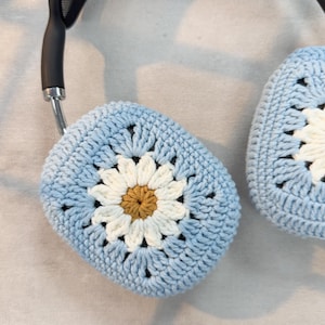 Puede incluir: Un par de fundas para auriculares de ganchillo azul con un dise&ntilde;o de margarita blanca. La margarita tiene un centro amarillo.