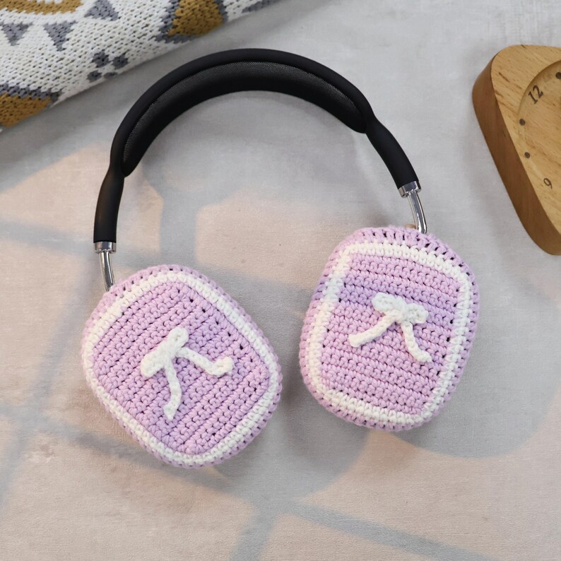 May include: Purple crocheted earmuffs with white trim and a white bow design. The earmuffs are attached to a black headband.