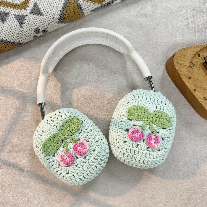 Pu&ograve; includere: Un paio di cuffie bianche con paraorecchie all'uncinetto. I paraorecchie sono di colore verde chiaro con disegni di ciliegie rosa e verdi.