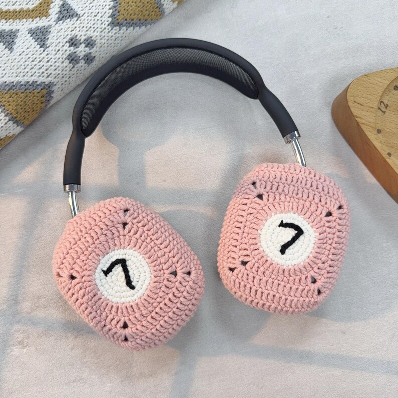 K&ouml;nnte beinhalten: Rosa geh&auml;kelte Ohrensch&uuml;tzer mit einem wei&szlig;en Kreis und der Zahl "7" auf jeder Ohrmuschel gestickt. Die Ohrensch&uuml;tzer sind an einem schwarzen Kopfband mit einem Netzfutter befestigt.