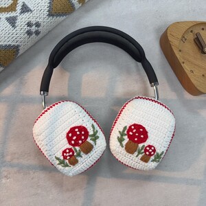 May include: White crocheted headphone covers with red and white mushroom embroidery. The covers are attached to a black headband.