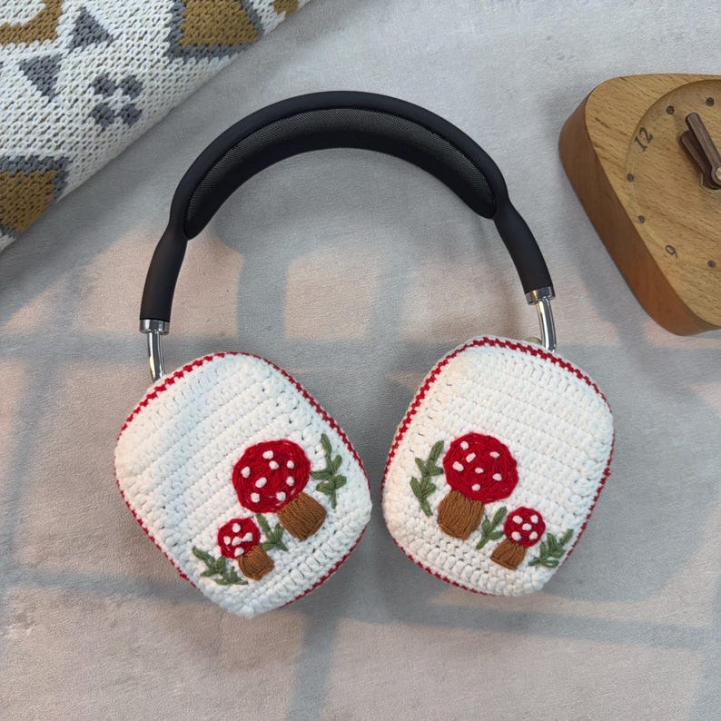 May include: White crocheted headphone covers with red and white mushroom embroidery. The covers are attached to a black headband.