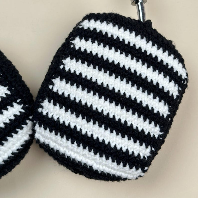 May include: Close-up of a pair of black and white striped crochet ear covers. The ear covers are made of a soft yarn and have a repeating pattern of black and white horizontal stripes. The ear covers are designed to fit over headphones.