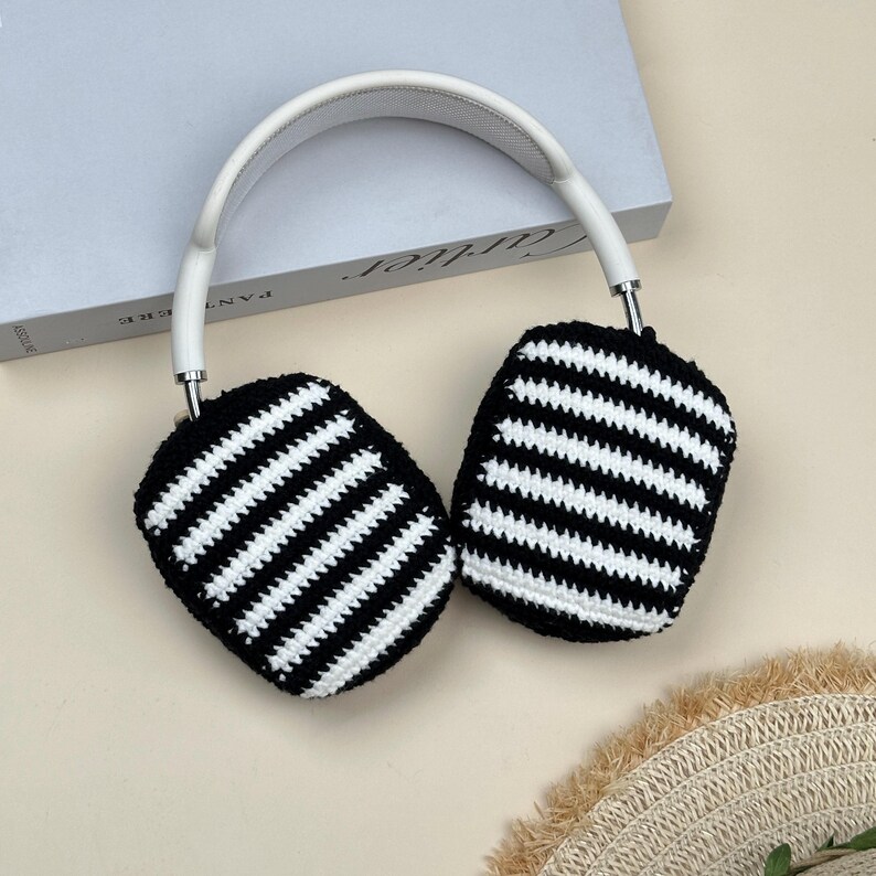 May include: White and black striped crochet headphone covers with a white headband. The ear covers are black with white horizontal stripes. The headband is white. The headphones are resting on a light beige surface.