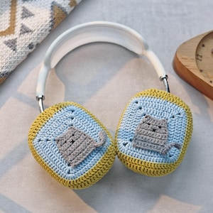 Pu&ograve; includere: Un paio di cuffie bianche con paraorecchie all'uncinetto. I paraorecchie sono blu, gialli e grigi con un design a forma di gatto.