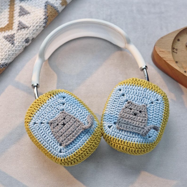 Pu&ograve; includere: Un paio di cuffie bianche con paraorecchie all'uncinetto. I paraorecchie sono blu, gialli e grigi con un design a forma di gatto.