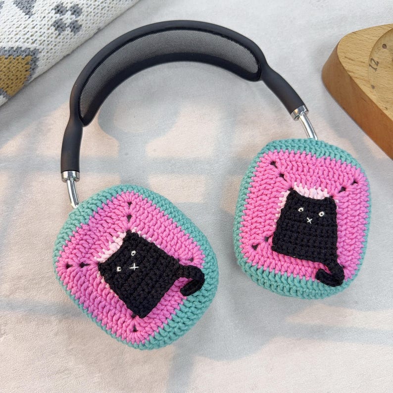 May include: A pair of black headphones with crocheted earmuffs. The earmuffs are pink and green with a black cat design.